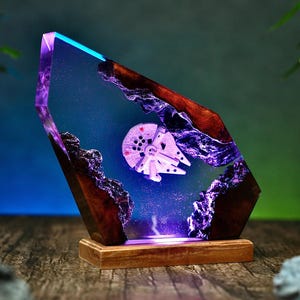 Pode incluir: Uma peça de arte em resina única com um modelo da Millennium Falcon. A obra de arte é encapsulada em resina transparente com um gradiente azul e roxo, acentuada por madeira escura e uma base de madeira. O modelo da nave espacial é iluminado.