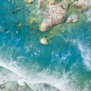 May include: Close-up of a resin art piece depicting a turquoise ocean with white foamy waves crashing on a sandy shore. The ocean floor is visible with small rocks and sand.