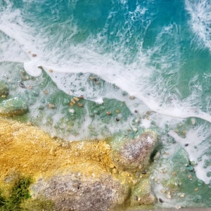 May include: Close-up of a resin art piece depicting a beach scene. The image shows a sandy shore with rocks and green foliage, meeting a turquoise blue ocean with white foamy waves.