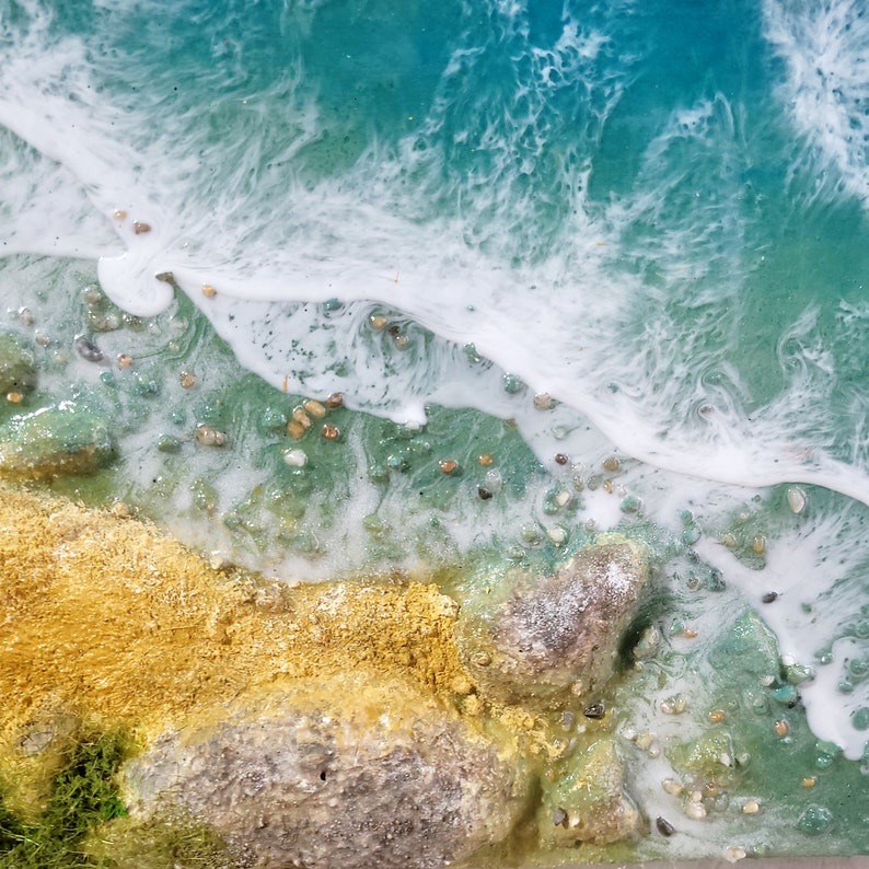May include: Close-up of a resin art piece depicting a beach scene. The image shows a sandy shore with rocks and green foliage, meeting a turquoise blue ocean with white foamy waves.