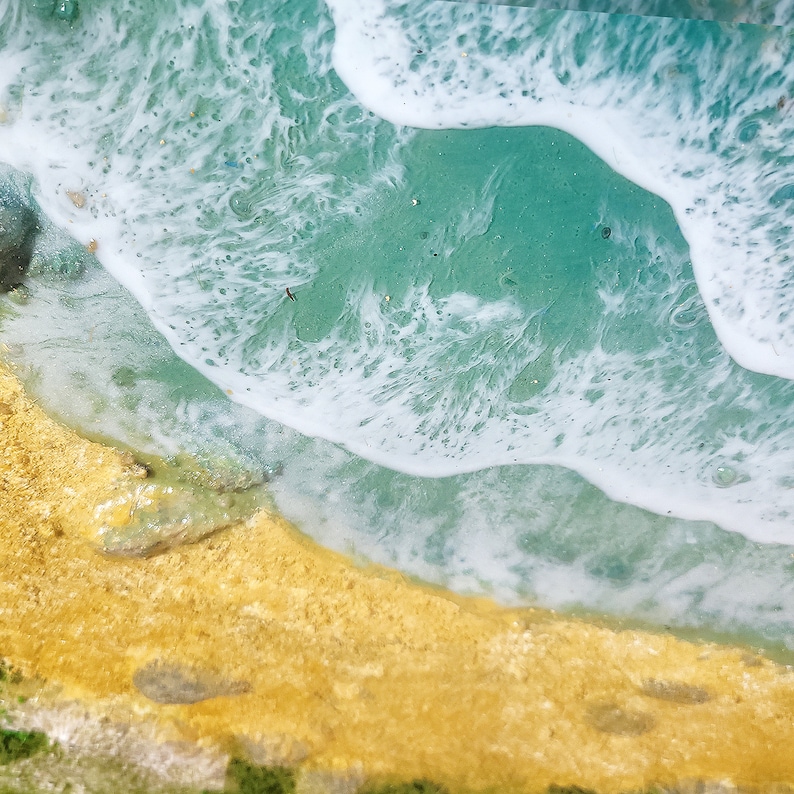 May include: A close-up of a resin art piece depicting a beach scene. The piece features a turquoise blue ocean with white foamy waves crashing on a golden sand beach.