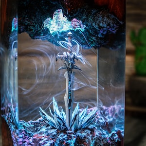 Puede incluir: Una espada plateada con mango negro est&aacute; encapsulada en resina transparente con un efecto de remolino azul y morado. La espada est&aacute; rodeada de un efecto de humo blanco y vaporoso.