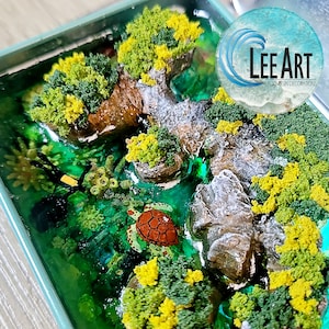 May include: A miniature resin art piece depicting a small island with a green and yellow mossy forest, a small waterfall, and a turtle swimming in the turquoise water. The art piece is framed in a light blue box with the text "LEE ART EPOXY RESIN DECORATIONS" on the side.