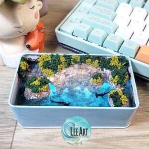 May include: A small, blue, rectangular box containing a resin scene. The scene depicts a small, rocky island with green foliage and a blue lagoon with two dark shapes that resemble fish. The box has a white label on the bottom with the text "LEE ART" and the words "YOUR OWN DECORATIONS".