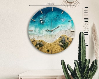 Puede incluir: Un reloj redondo de resina con una escena de playa. La esfera del reloj es azul y verde con olas blancas y arena. Las manecillas del reloj son doradas. El reloj tiene un diámetro de 40 cm.