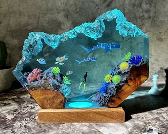 Könnte beinhalten: Eine Harzskulptur, die eine Unterwasserszene in Blau-, Grün- und Brauntönen darstellt. Die Skulptur zeigt einen Taucher, zwei Haie, eine Schildkröte und Korallen. Die Skulptur wird von unten beleuchtet, wodurch ein leuchtender Effekt entsteht.