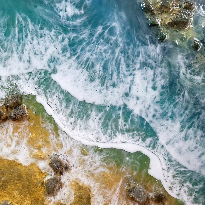 Pode incluir: Um close-up de uma obra de arte em resina que retrata uma cena de praia. A imagem mostra um oceano azul turquesa com ondas brancas espumosas quebrando em uma costa arenosa com rochas marrons.