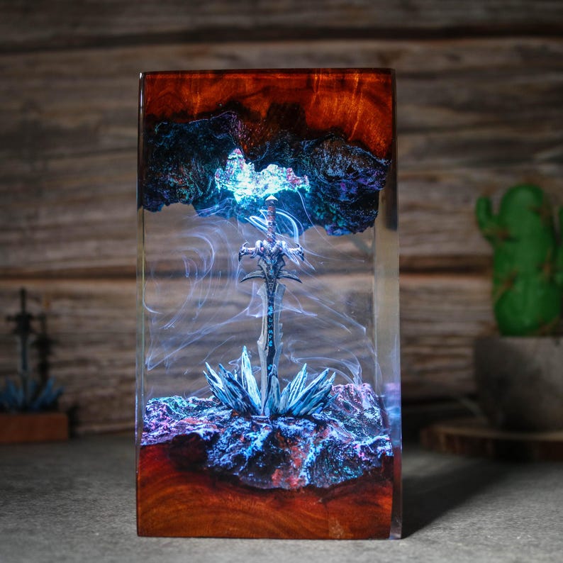 Puede incluir: Una espada plateada est&aacute; encapsulada en resina transparente con remolinos azules y morados. La resina est&aacute; colocada en una base de madera con un acabado marr&oacute;n oscuro.