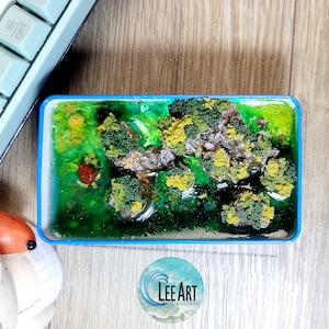 以下が含まれることがあります： 青い枠のある小さな長方形のレジンアート作品。作品は、緑と黄色の葉、小さな茶色の亀、透明なレジン川のあるミニチュアの森のシーンを描いています。