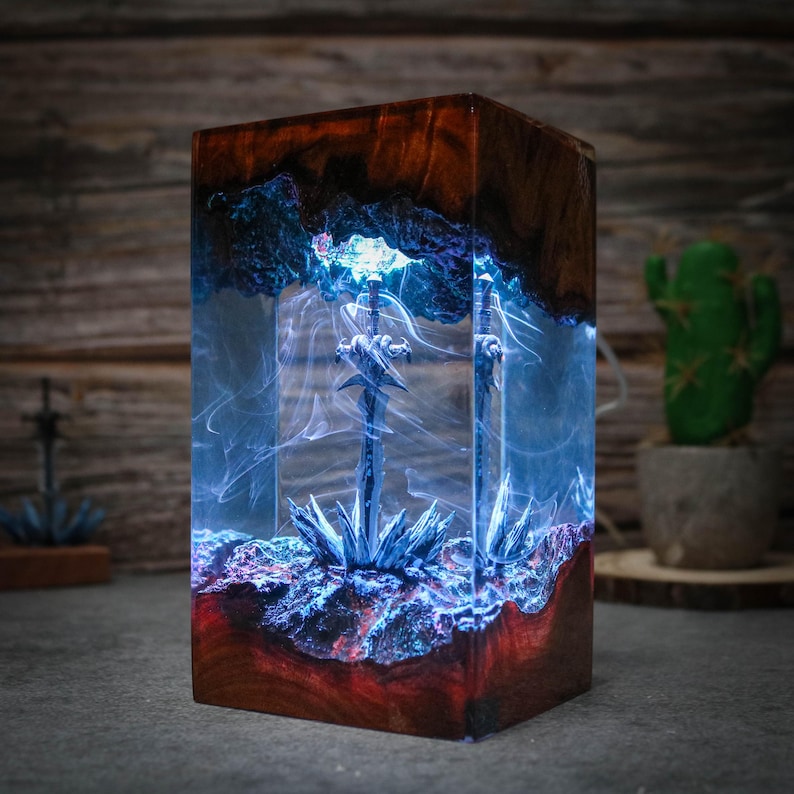 Puede incluir: Una base de l&aacute;mpara de madera con un bloque de resina transparente que contiene dos espadas plateadas y patrones azules y blancos en espiral. La base es de madera marr&oacute;n oscura con vetas rojas y azules.