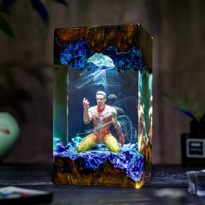 Può includere: Scultura rettangolare in resina con una figura di Iron Man in ginocchio. La scultura è racchiusa in una resina trasparente con accenti blu e oro, creando un effetto a strati. La figura è dettagliata con un'armatura rossa e oro.