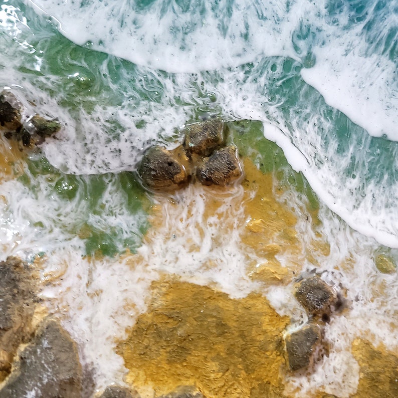Pode incluir: Close-up de uma obra de arte em resina que representa uma cena de praia com ondas brancas espumosas quebrando em uma costa arenosa com rochas verdes e marrons.