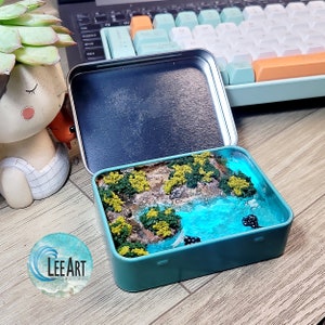 May include: A small, teal-coloured tin box with a clear resin scene inside. The scene depicts a miniature landscape with a blue river, green trees, and brown rocks. The tin box is sitting on a wooden surface.