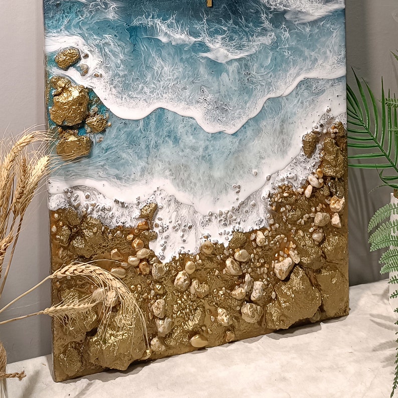 K&ouml;nnte beinhalten: Abstrakte Harz-Kunstmalerei, die eine Strandszene mit blauen und wei&szlig;en Wellen darstellt, die an einem Ufer aus goldenen und wei&szlig;en Steinen brechen.