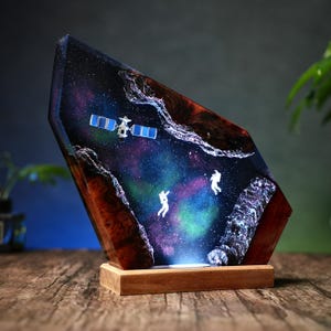 May include: A unique resin art piece depicting a space scene with astronauts and a satellite. The artwork features a colorful galaxy design with shades of blue, purple, and green, set within a wooden base. The piece is illuminated from below.