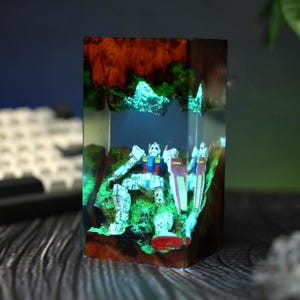Puede incluir: Una pieza de arte de resina rectangular con una figura de robot detallada dentro de un paisaje en miniatura. La obra de arte muestra una paleta de colores vibrantes con acentos verdes, rojos, azules y blancos, y un efecto brillante.