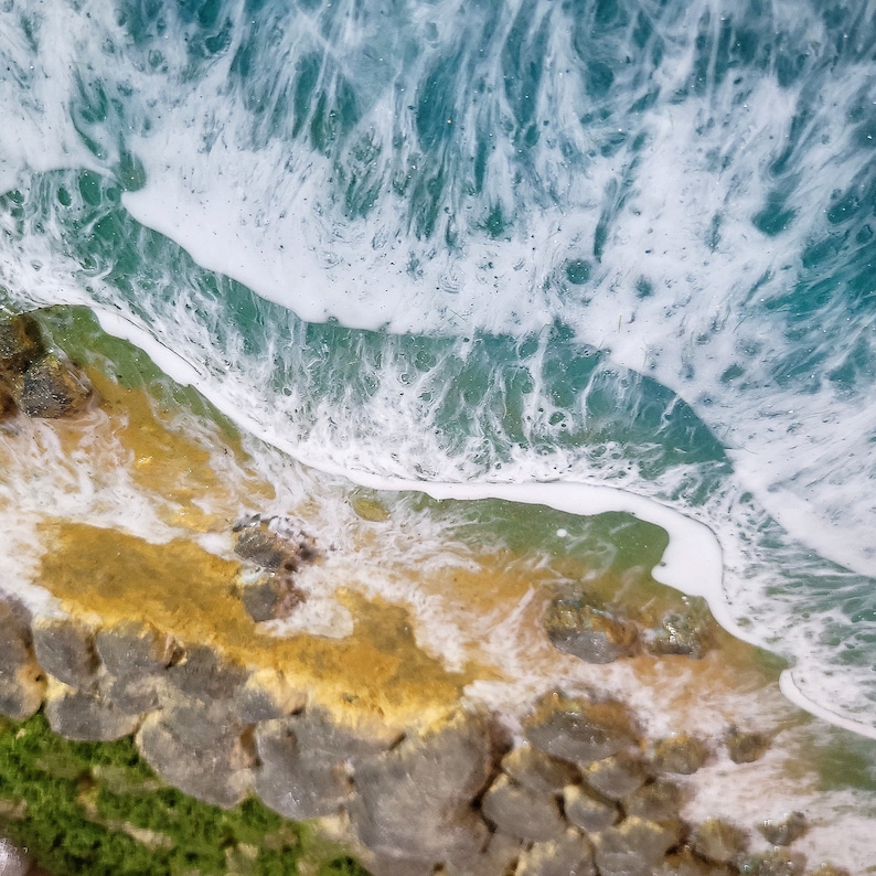 Pode incluir: Close-up de uma obra de arte em resina que representa uma onda quebrando em uma costa rochosa. A onda &eacute; branca com tons azuis e verdes, e a costa &eacute; de cor amarela arenosa com rochas marrons.
