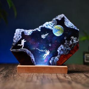 Pode incluir: Escultura decorativa em resina retratando uma cena espacial com astronautas, uma nave espacial e um planeta. A escultura está colocada sobre uma base de madeira. O fundo apresenta um design de galáxia colorido.