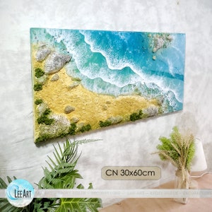May include: A blue and white epoxy resin art piece depicting an aerial view of a beach with waves crashing on the shore. The beach is made of sand and has small rocks and green moss. The art piece is 30 inches by 60 inches.