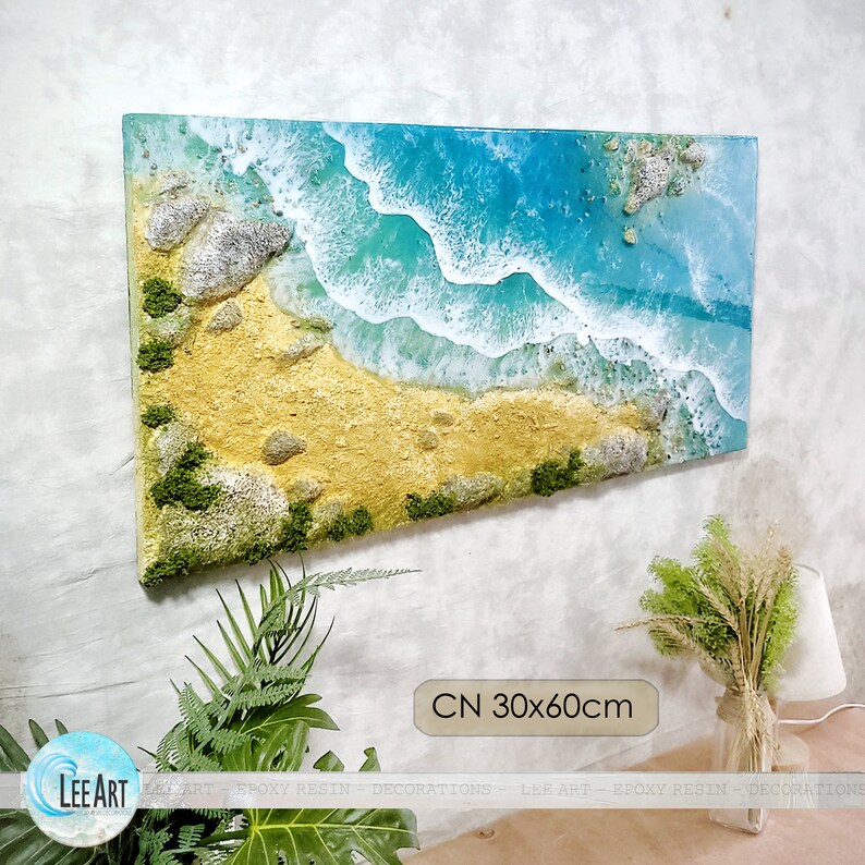 May include: A blue and white epoxy resin art piece depicting an aerial view of a beach with waves crashing on the shore. The beach is made of sand and has small rocks and green moss. The art piece is 30 inches by 60 inches.