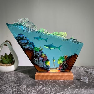 May include: A resin sculpture depicting an underwater scene with two sharks, coral, and seaweed. The sculpture is clear and has a blue and green tint. It is mounted on a wooden base.