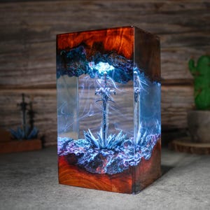 Puede incluir: Una espada azul brillante encapsulada en resina transparente con un fondo azul oscuro y negro en espiral. La resina est&aacute; colocada en una base de madera con un acabado marr&oacute;n rojizo.