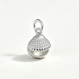 May include: A polished silver charm shaped like a small, open seashell. The exterior of the shell features a textured, beaded pattern, while the interior is smooth. A small, circular silver jump ring is attached to the top for hanging.