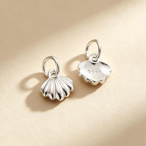 Peut inclure: Deux petites breloques en forme de coquillage argenté, chacune dotée d'un anneau de jonction circulaire sur le dessus. L'une présente une texture nervurée en éventail, tandis que l'autre montre un dos lisse avec l'inscription 925 gravée.