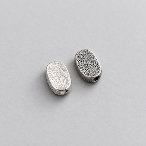 Peut inclure: Deux perles de forme ovale avec une surface texturée et irrégulière. Une perle est d'un ton argenté brillant et poli, tandis que l'autre est d'une couleur argentée oxydée plus foncée. Chaque perle présente un petit trou visible sur le côté.