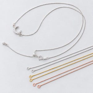 May include: A silver-coloured necklace featuring a crescent moon pendant encrusted with small clear stones, accompanied by several additional thin box chains in silver, gold, and rose gold tones, each finished with a small circular end ring.