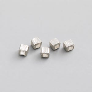 Può includere: Cinque piccole perline a forma di cubo color argento con una finitura metallica spazzolata. Ogni perlina presenta un foro quadrato centrale che attraversa il centro, mostrando un design geometrico uniforme con bordi leggermente arrotondati e una texture opaca.