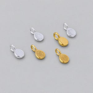 May include: A collection of six small, oval-shaped jewelry charms, each featuring a circular jump ring at the top. Three of the charms have a polished silver-tone finish, while the other three have a vibrant gold-tone finish with textured surfaces.