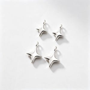 Puede incluir: Cuatro dijes plateados pulidos en forma de estrella, cada uno con un diseño suave y redondeado de cuatro puntas con un pequeño lazo circular en la parte superior para sujetarlos. Los dijes están dispuestos en un grupo suelto.
