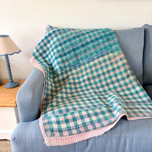 May include: A knitted throw blanket featuring a chequered pattern in shades of teal, light blue, and soft pink. The blanket has a visible textured stitch throughout and a solid light pink border along the edges, draped over a blue sofa.