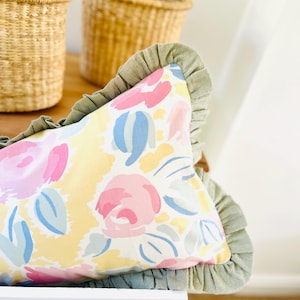 Puede incluir: Un cojín decorativo con un estampado floral en tonos rosa, azul y amarillo. El cojín tiene un borde verde con volantes y está colocado sobre una superficie blanca. Dos cestas tejidas son visibles en el fondo.