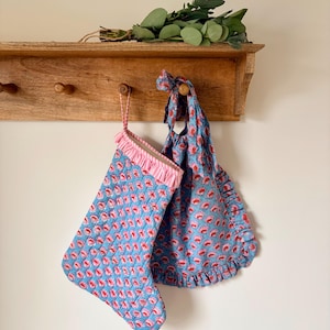 May include: A quilted Christmas stocking and a matching fabric bag, both featuring a blue background with a repeating pink and red floral pattern. The stocking has a pink and white striped ruffled cuff, and the bag has a ruffled edge.