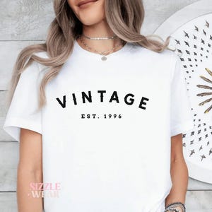 以下が含まれることがあります： 「VINTAGE」の文字が黒い太字で「EST. 1996」の上に書かれた白いTシャツ。Tシャツは半袖で、クルーネックです。Tシャツは人が着ています。