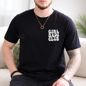 May include: Black crew neck t-shirt with the white text "GIRL DADS CLUB" on the right chest. The shirt also has the text "SIZZLE WEAR" on the left sleeve. A gold necklace with a circular pendant is visible.