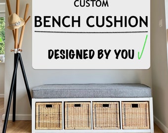 May include: A white rectangular sign with black text reading CUSTOM BENCH CUSHION DESIGNED BY YOU with a green checkmark. Below, a white bench features a grey upholstered cushion and four woven wicker storage baskets underneath.