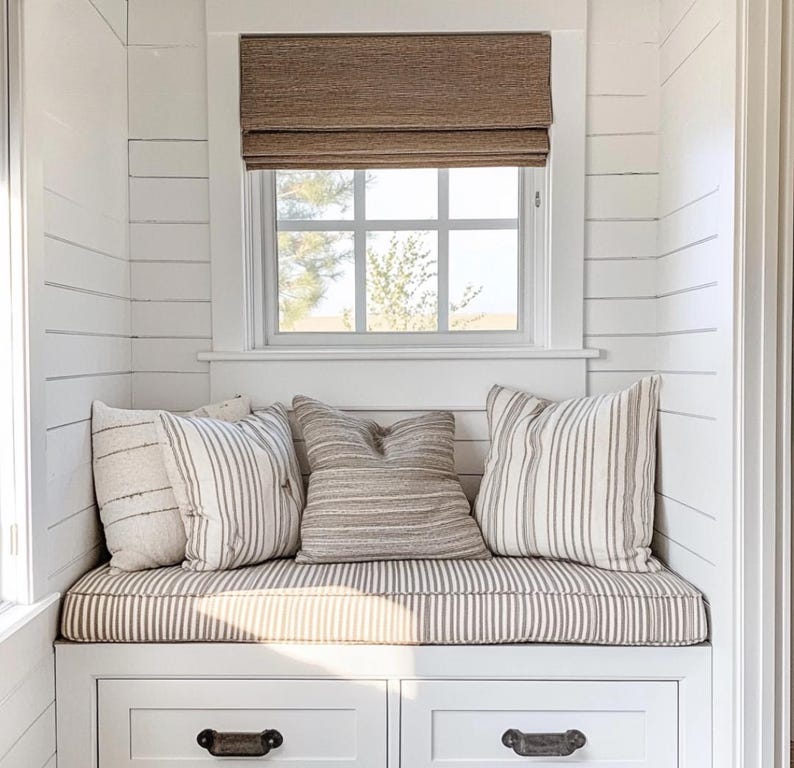 May include: A white built-in window seat with two drawers featuring dark metal pulls. The seat cushion is striped in beige and white. Three decorative pillows are arranged on the seat, including two striped pillows and one solid textured beige pillow.