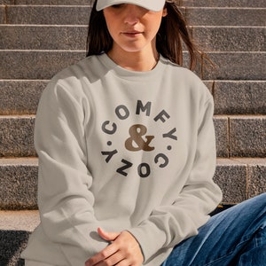 May include: A light gray sweatshirt with a black circle graphic that says "COMFY & COZY" in a circular design. The sweatshirt is being worn by a person wearing a white baseball cap and blue jeans.