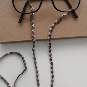 May include: Black eyeglasses with a decorative eyeglass chain. The chain is dark green with pink bead accents. The glasses are resting on a brown surface.