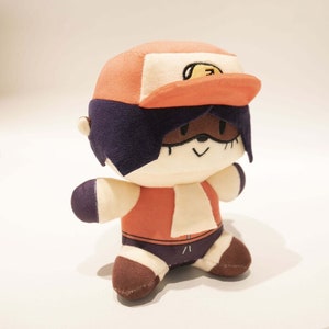 Puede incluir: Un juguete de peluche de un personaje de dibujos animados que lleva una gorra de béisbol roja y blanca con una cara sonriente amarilla. El personaje tiene piel marrón, cabello negro y una camisa blanca con un chaleco rojo. El juguete está sentado sobre una superficie blanca.