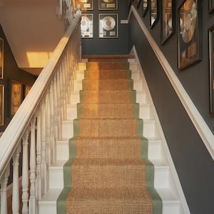 May include: A staircase with white steps and balustrades, a natural fibre runner with a green border, and dark grey walls. Framed gold record awards adorn the walls. The stairs are illuminated by warm sunlight.