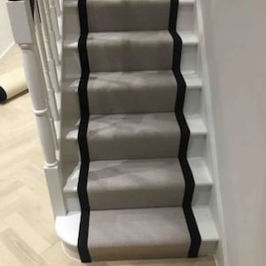 May include: A set of stairs with a light grey carpet runner and black trim along the edges. The stairs are carpeted in a light grey colour and have a black border along the edges. The stairs are made of wood and have a white railing.