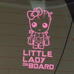 Op de afbeelding: Roze vinyl sticker van een cartoonfiguur met een strik op zijn hoofd. Het figuur staat met zijn armen langs zijn zijde. De tekst "LITTLE LADY ON BOARD" staat onder het figuur.