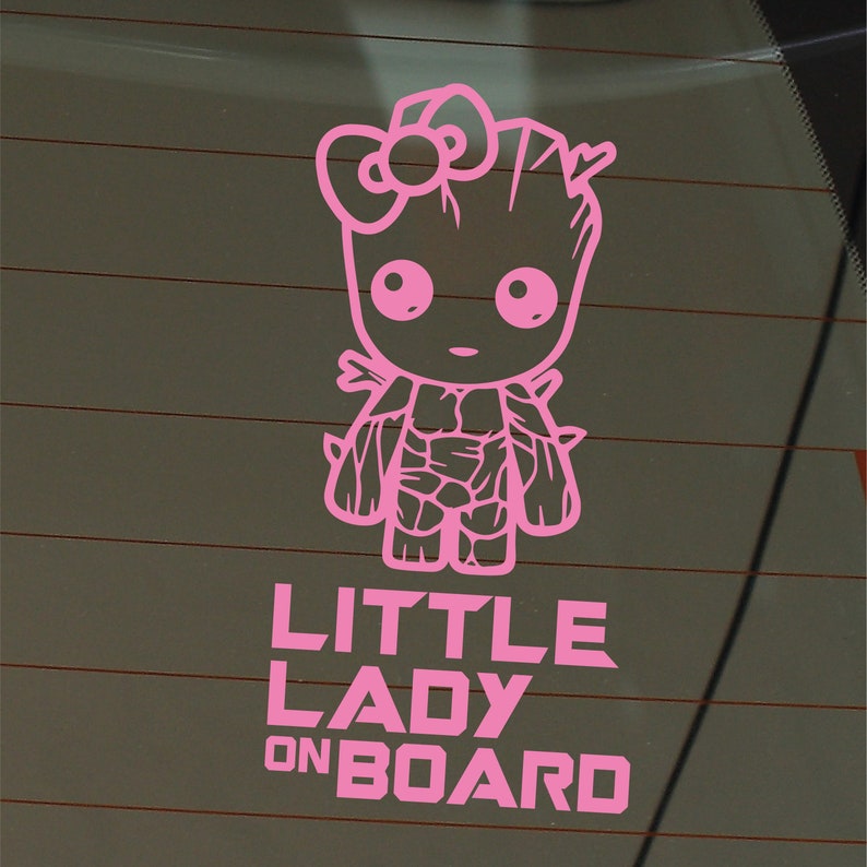 Op de afbeelding: Roze vinyl sticker van een cartoonfiguur met een strik op zijn hoofd. Het figuur staat met zijn armen langs zijn zijde. De tekst "LITTLE LADY ON BOARD" staat onder het figuur.