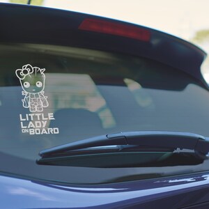 Op de afbeelding: Een witte vinyl sticker met een cartoonfiguur en de tekst "LITTLE LADY ON BOARD". De sticker zit op de achterruit van een blauwe auto.