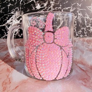 May include: A clear glass mug with a pink rhinestone pumpkin design. The pumpkin has a bow and stem, all covered in sparkling rhinestones. The mug has a handle and is set against a marble background.