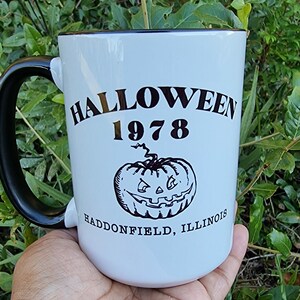 May include: White ceramic mug with a black handle and rim. The mug features the text "HALLOWEEN 1978" above a jack-o'-lantern graphic, with "HADDONFIELD, ILLINOIS" below. The mug is held in a hand against a backdrop of green foliage.
